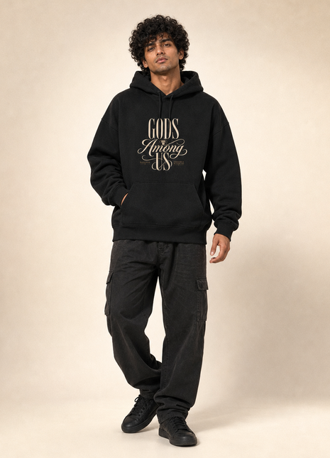 Indian male model wearing PAXBURY Gods Among Us black unisex UH32 hoodie with original-style cream GODS Among US typography centred above front pouch