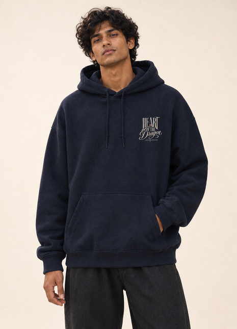 Indian male model front view wearing PAXBURY Heart of the Dragon navy blue UH32 unisex oversized hoodie with exact-style small ornate cream print on wearer's left chest above kangaroo pocket
