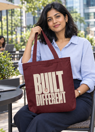 Built Different maroon canvas tote, large type front, young Indian office woman seated at outdoor cafe