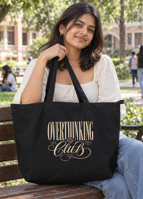 Overthinking Club large black zip tote: elegant front graphic, Indian college student seated on a campus bench