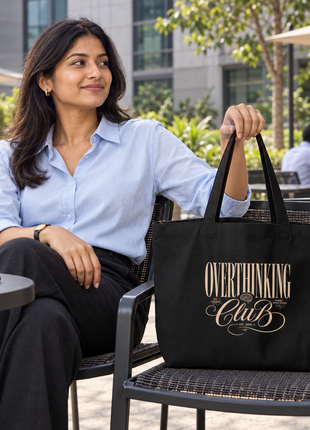 Overthinking Club black large tote: elegant front artwork, Indian young professional seated outside office café