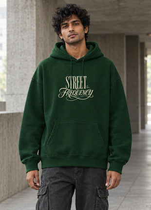 Indian male model wearing Street Frequency forest-green oversized hoodie, cream STREET Frequency vintage typography on chest